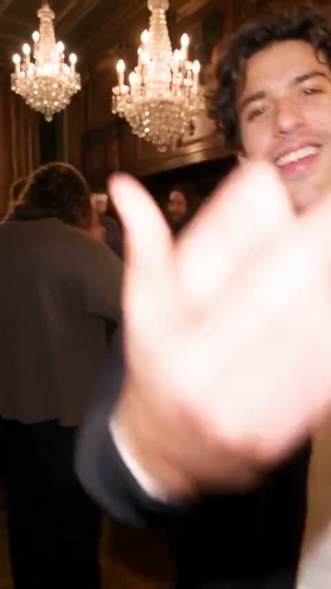 A guy in a bomber jacket, cup up, in a gilded ballroom.